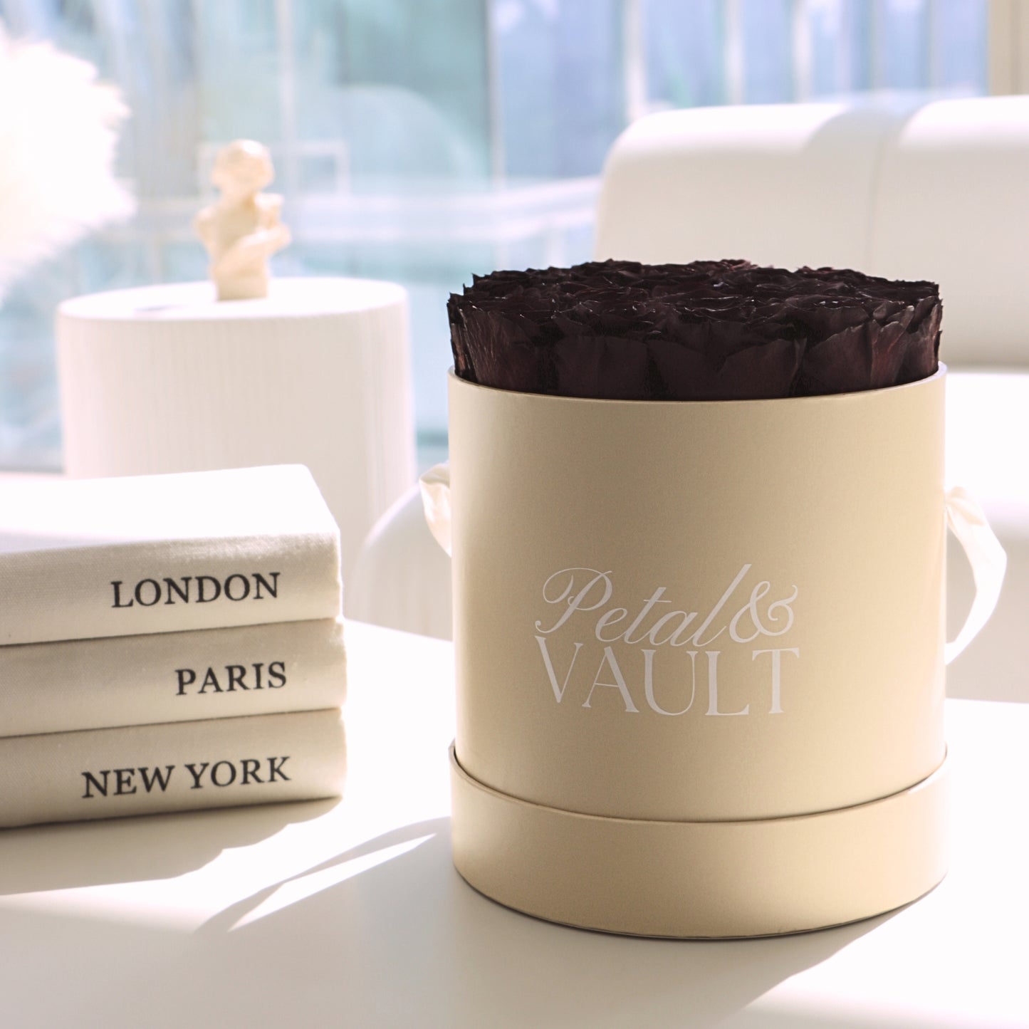 Beige hat box container with 'Petal & Vault' branding, burgundy preserved rose inside, books labeled 'London', 'Paris', 'New York' in the background.