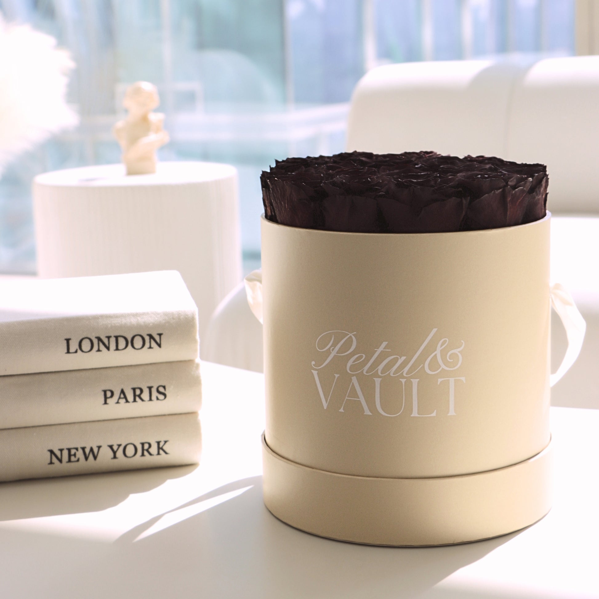 Beige hat box container with 'Petal & Vault' branding, burgundy preserved rose inside, books labeled 'London', 'Paris', 'New York' in the background.