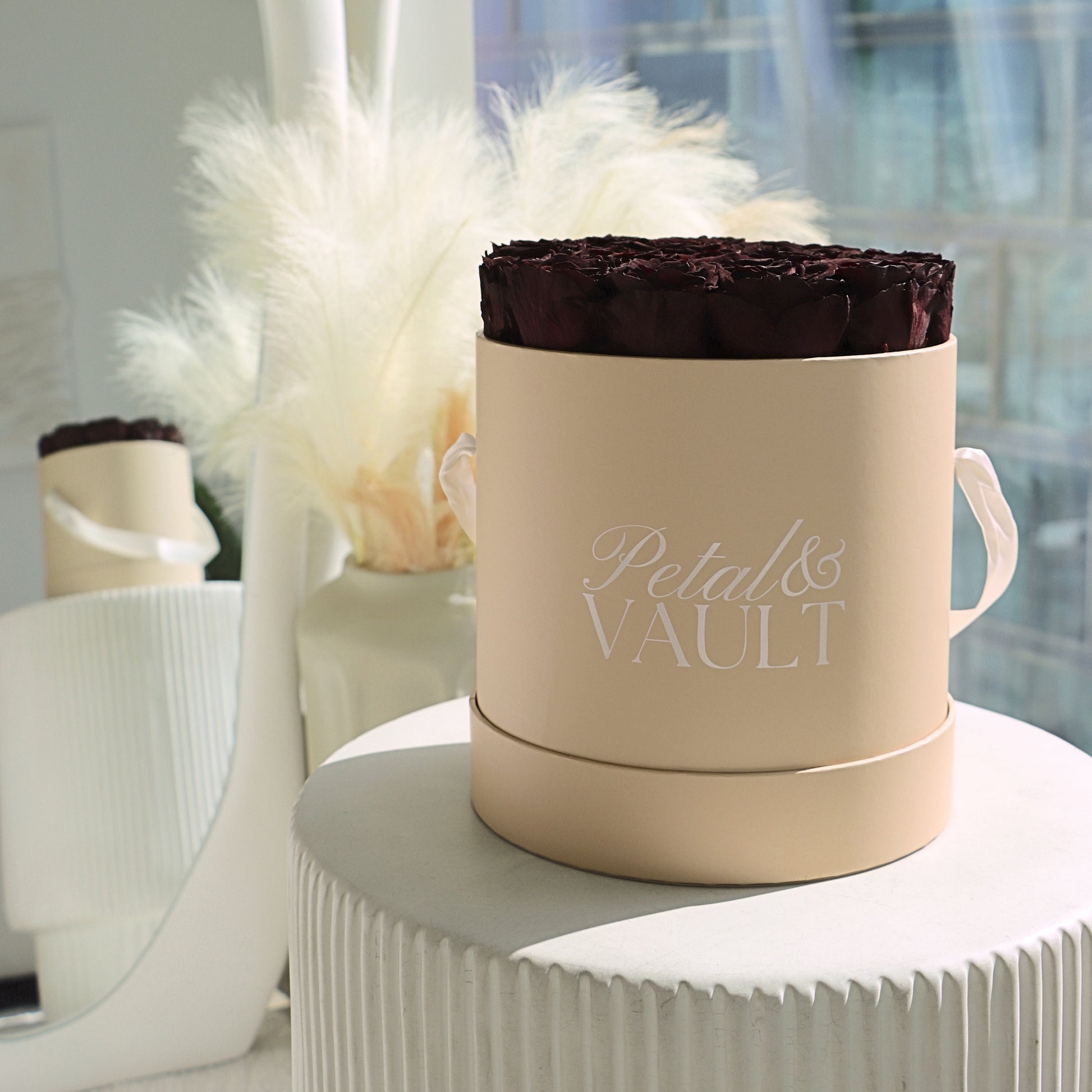 Beige round hat box container with a dark red rose arrangement labeled 'Petal & Vault' on a white surface.