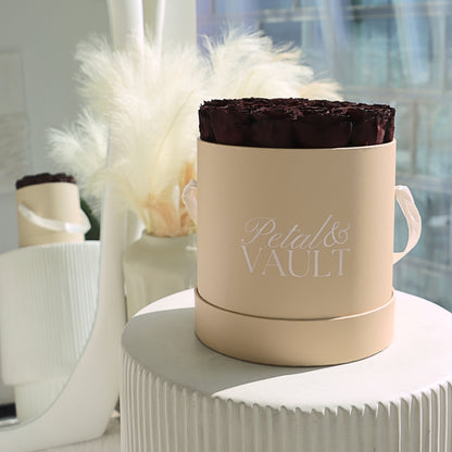 Beige round hat box container with a dark red rose arrangement labeled 'Petal & Vault' on a white surface.