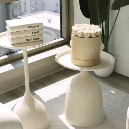 White side table with a decorative rose hat box and books in a room with large windows.