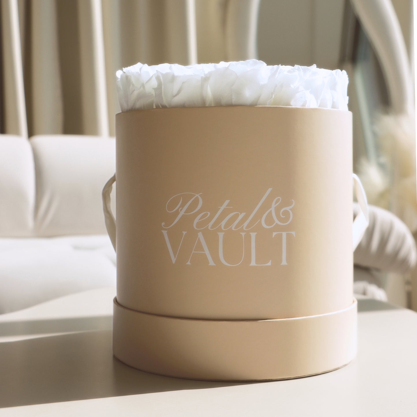 Beige cylindrical hat box with 'Petal & Vault' branding containing white roses on a coffee table