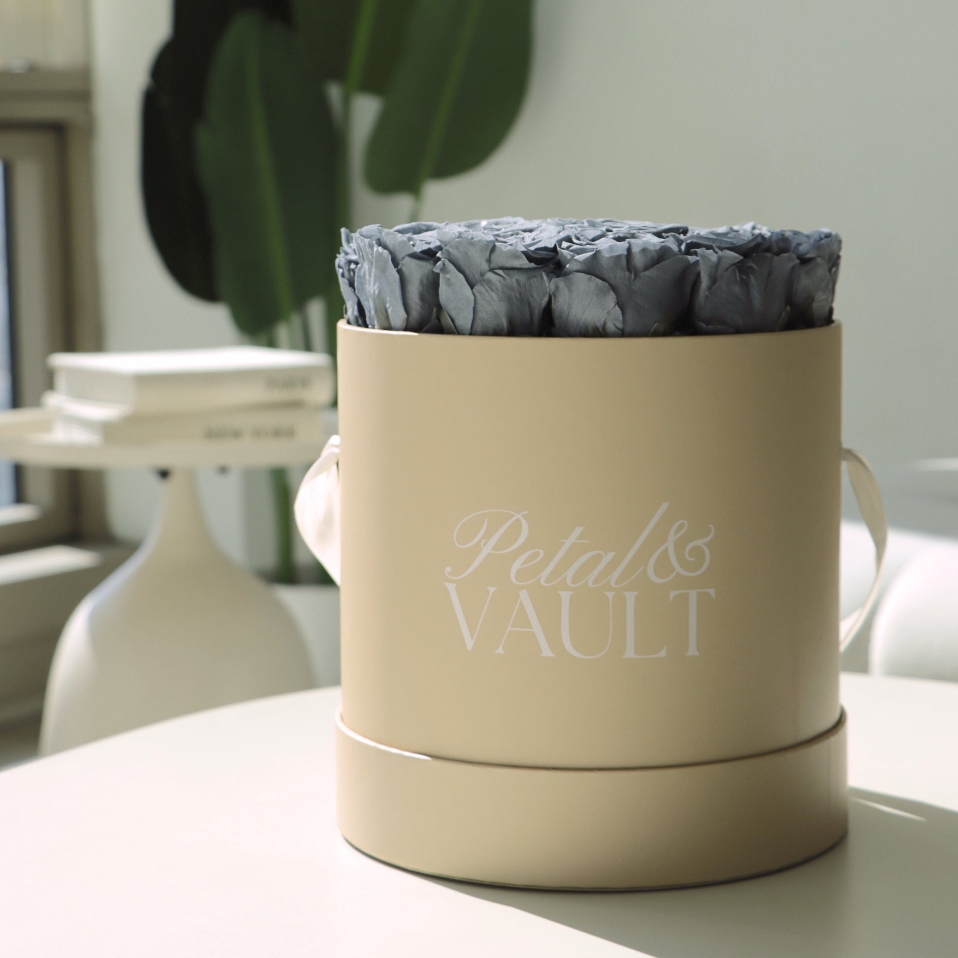 Beige cylindrical hat box container with 'Petal & Vault' branding, filled with blue flowers, on a light coffee table surface.