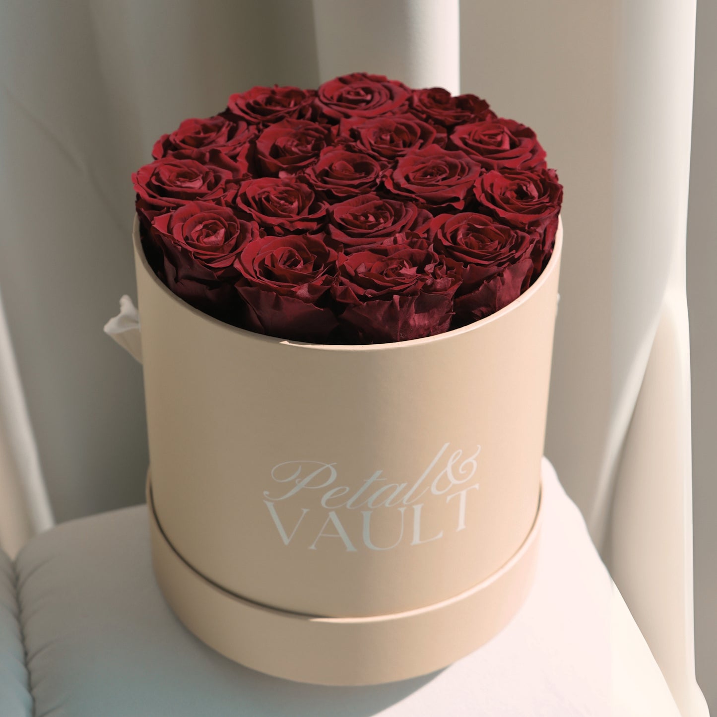 Beige round hat box with red roses labeled 'Petal & Vault' on n ivory couch surface.