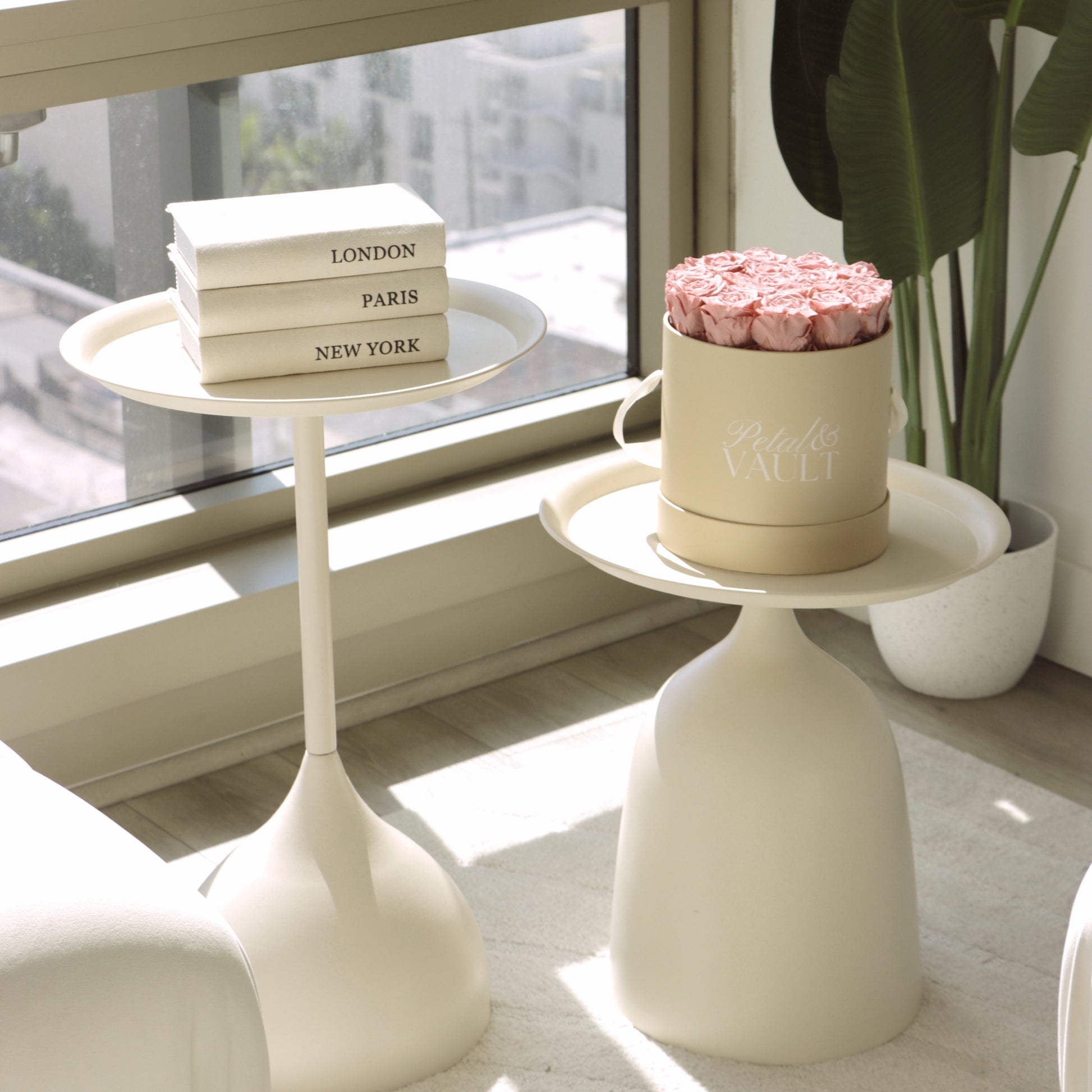 Two small white side tables with a hat box  filled with dusty pink roses and books with  floor to ceiling windows in the background
